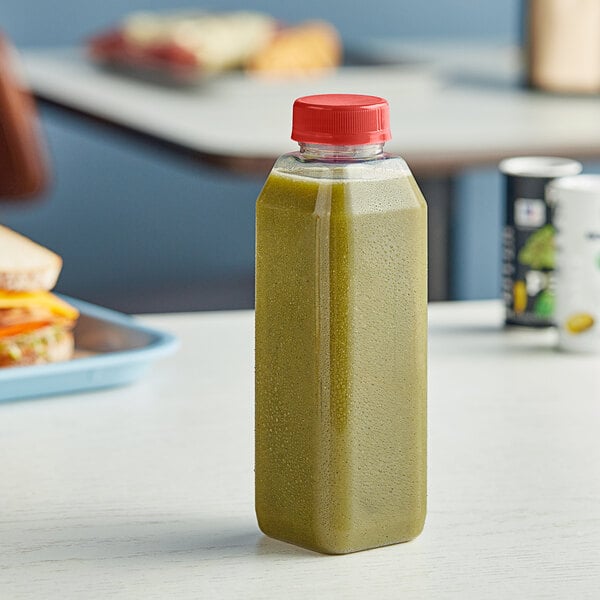 A 16 oz. tall square clear rPET juice bottle with green liquid and a red cap on a table.