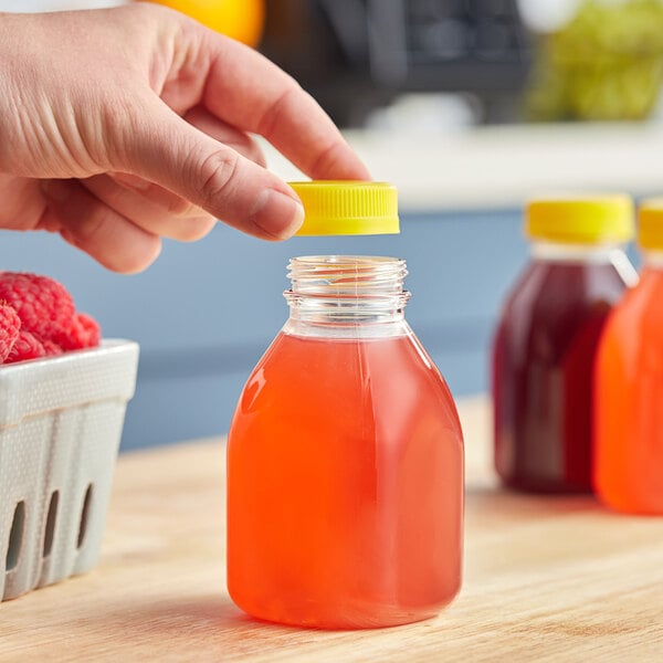 A hand holding a Square Clear PET juice bottle with a yellow lid filled with orange liquid.
