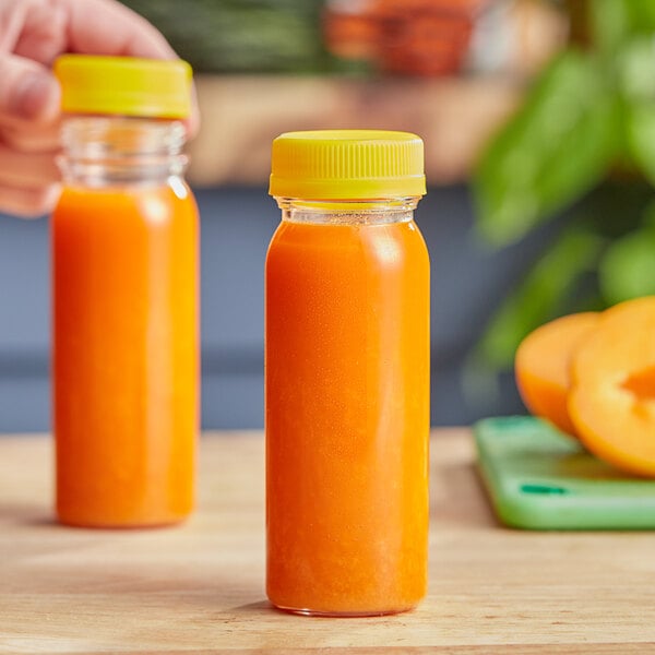 Two 4 oz. round PET clear juice bottles with yellow lids filled with orange liquid on a counter.
