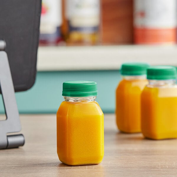 Square PET clear energy bottles with green lids filled with orange juice on a counter.