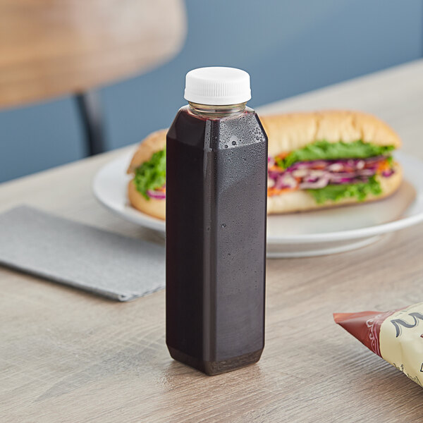 A tall square clear rPET juice bottle with a white lid next to a sandwich.