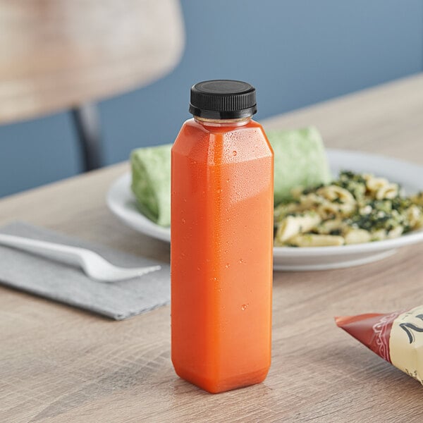 A 12 oz. tall square rPET clear juice bottle with black lid filled with orange juice on a table next to a plate of food.