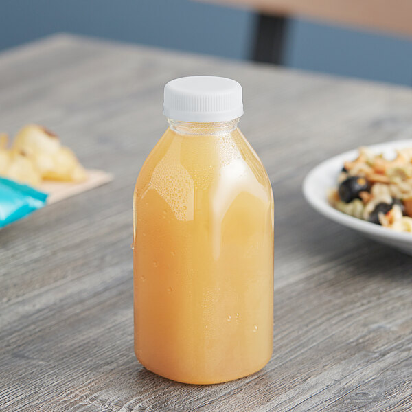 A 12 oz. Square Milkman PET clear juice bottle with a white lid on a table next to a bowl of fruit.