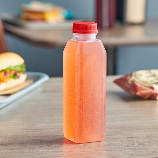 A 16 oz. tall square PET clear juice bottle with a red cap filled with pink juice on a counter.