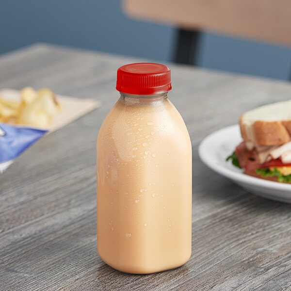 A 12 oz. square rPET clear juice bottle with a red lid filled with liquid on a table next to a sandwich.
