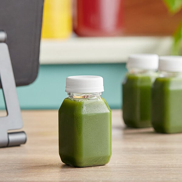 Three 4 oz. square PET clear energy juice bottles with white lids filled with green juice sit on a table.