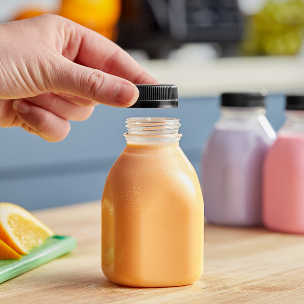 A hand holding a Square Milkman clear juice bottle with black lid filled with orange liquid.