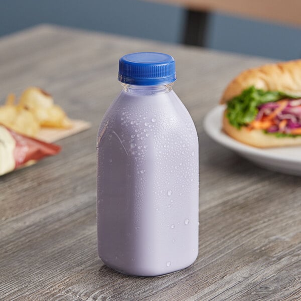 A Square Clear Milkman juice bottle with a blue lid on a table next to a sandwich.