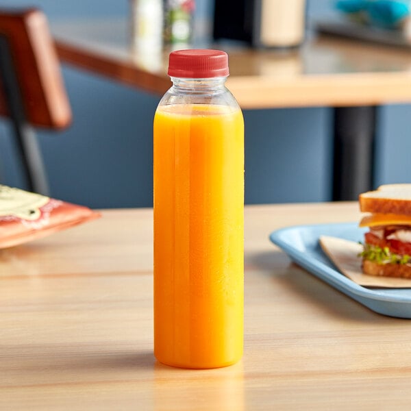 A 16 oz. round clear PET juice bottle with a red lid filled with orange juice on a table.