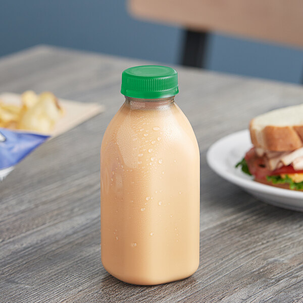 A 12 oz. square clear rPET juice bottle with a green cap on a table next to a plate of food.