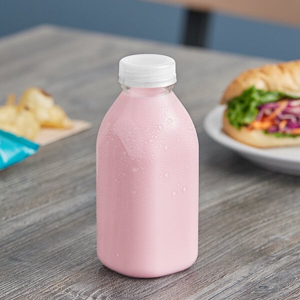 A clear Square Milkman juice bottle with pink liquid and a white lid on a table with a sandwich.