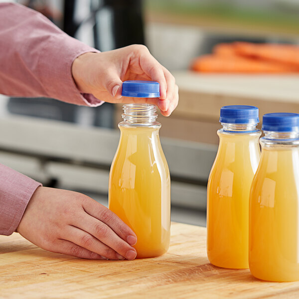 A hand pouring orange juice into a 12 oz. round clear carafe juice bottle with a blue lid.