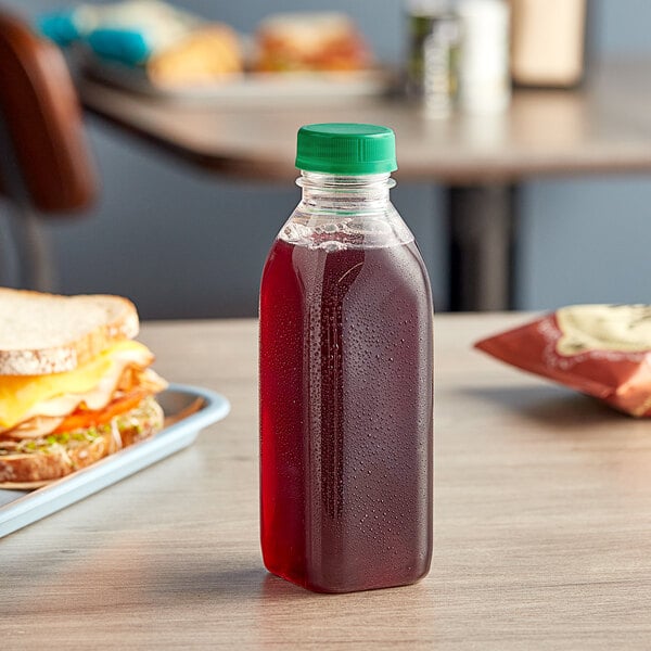 A 16 oz. Square Milkman PET Clear juice bottle with a dark green lid on a table next to a sandwich.