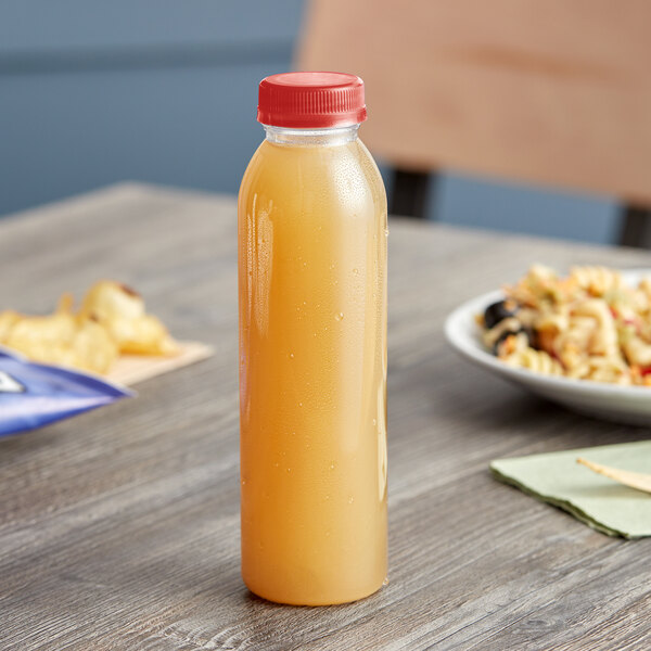 A round clear juice bottle with red lid on a table next to a plate of food.