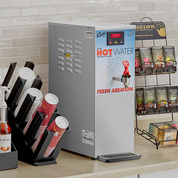 A Curtis dual voltage hot water dispenser on a counter with paper cups.