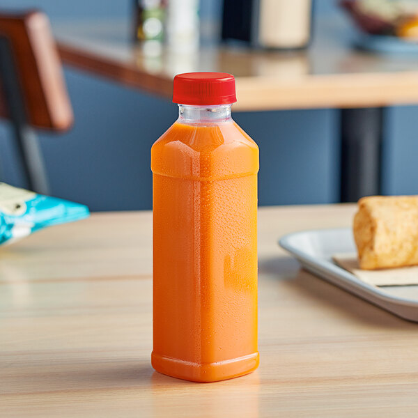 A 16 oz. Square PET clear juice bottle with a red lid and recessed label panel filled with orange juice on a table.