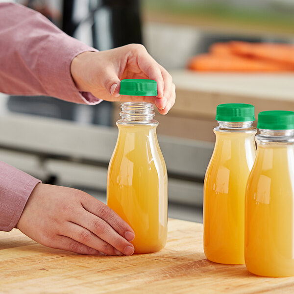 A person pouring orange juice into a round clear carafe with a green lid.