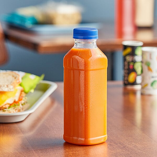 A 14.5 oz. square PET clear juice bottle with a blue lid on a table next to a sandwich.