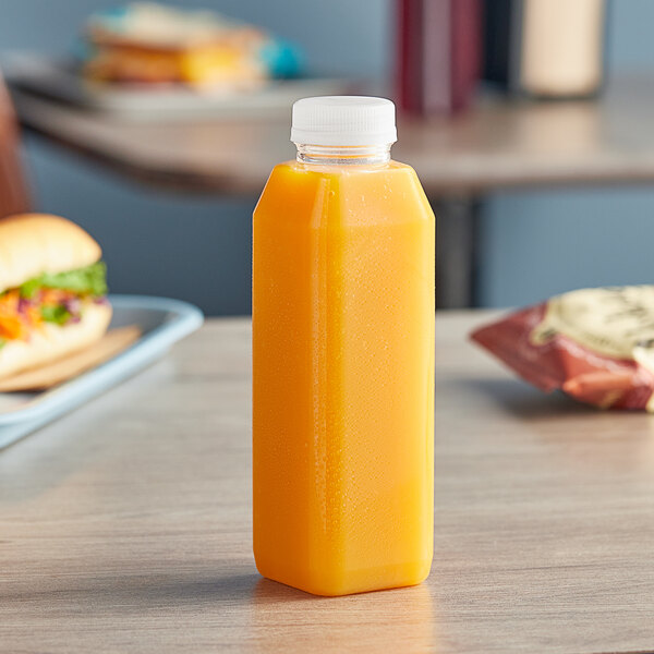 A 16 oz. tall square PET clear juice bottle with clear lid on a table next to a sandwich.