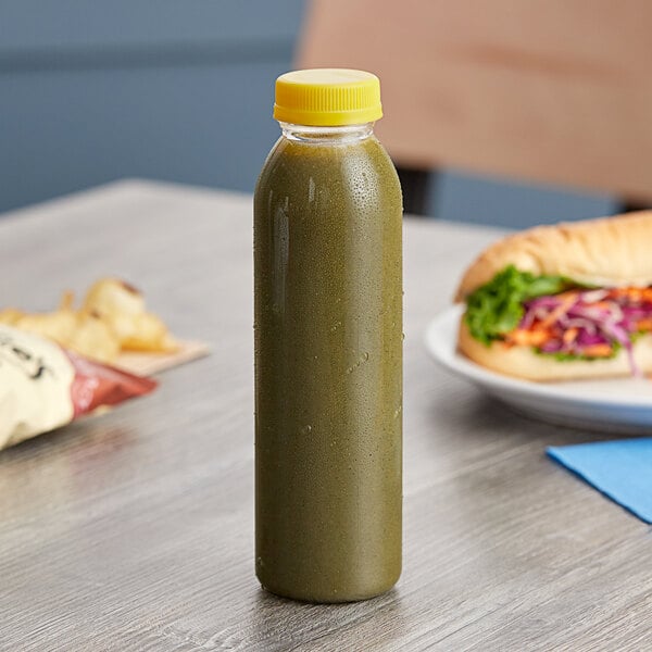 A clear round rPET juice bottle with a yellow cap on a table next to a sandwich.