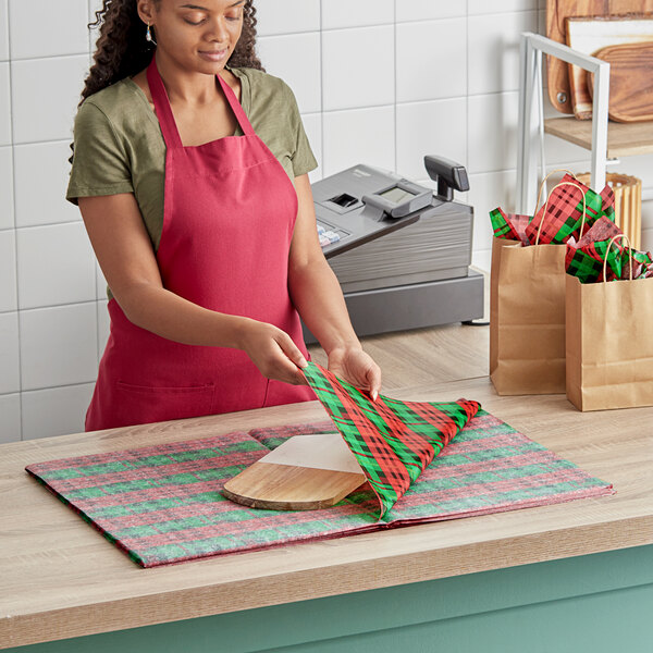 A woman in a red apron using SatinWrap red and green plaid tissue paper to wrap a brown bag.