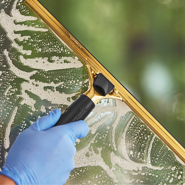 A hand wearing blue gloves using an Ettore Master black brass squeegee handle with a rubber grip to clean a window.