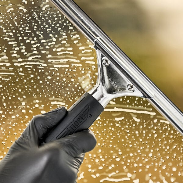 A gloved hand using an Ettore Master stainless steel squeegee with a black rubber grip to clean a window.