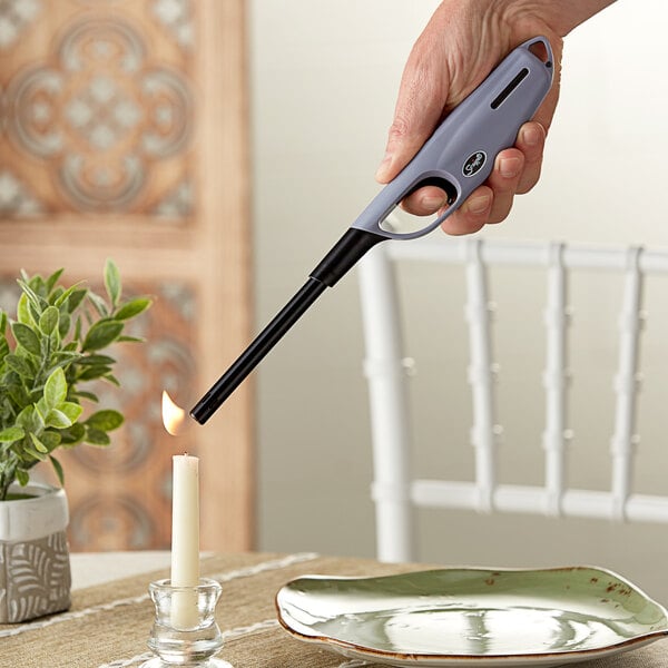 A hand holding a Scripto Aim 'N Flame Max Multi-Purpose Lighter over a candle in a glass holder.