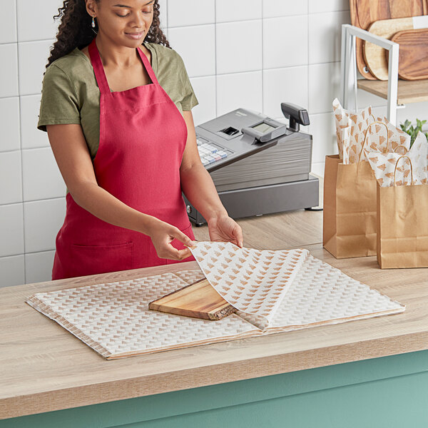 A woman in a red apron using SatinWrap gold pearl trees tissue paper sheets to wrap a product.