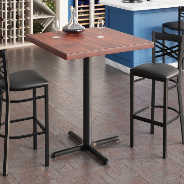 A Lancaster Table & Seating butcher block bar table with a mahogany finish and cast iron cross base plate with two wine glasses on it.