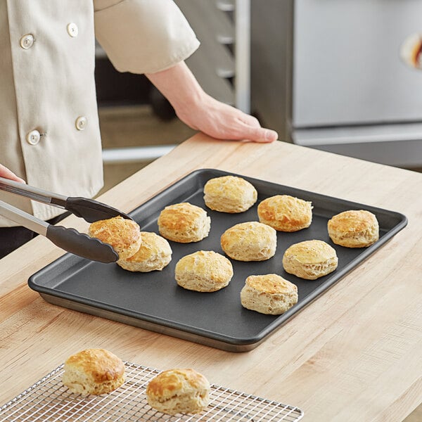 A person using tongs to pick up biscuits from a Baker's Lane non-stick sheet pan.