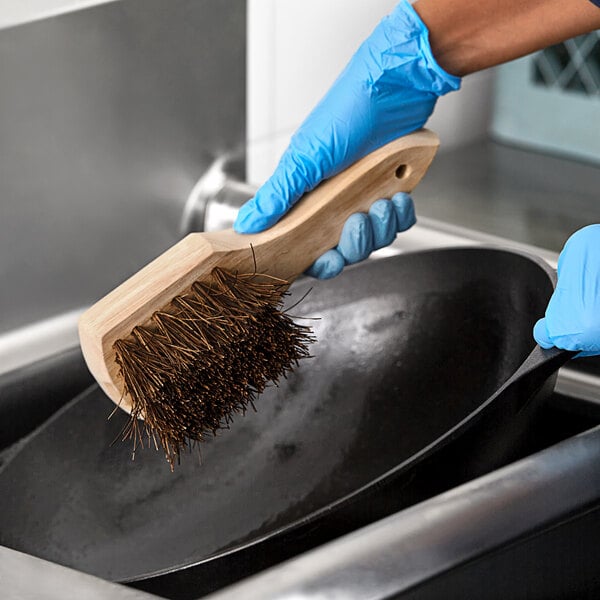 A person in blue gloves cleaning a frying pan with a Lavex Palmyra bristled wok brush.