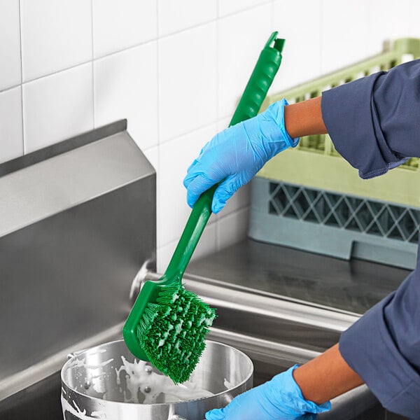 A person in blue gloves using a green Lavex floating pot scrub brush to clean a sink.