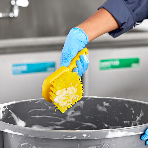 A hand in a blue glove using a Lavex yellow floating utility brush to clean a garbage can over a bucket of soapy water.