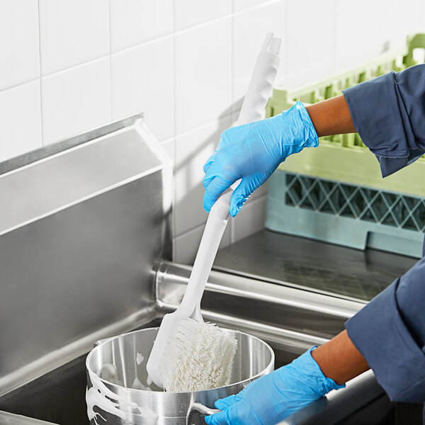 A person wearing blue gloves using a white Lavex pot scrub brush to stir food in a pot.