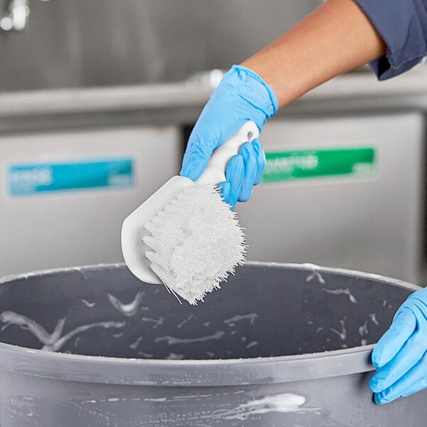 A person in blue gloves using a Lavex white floating utility brush to clean a large container.