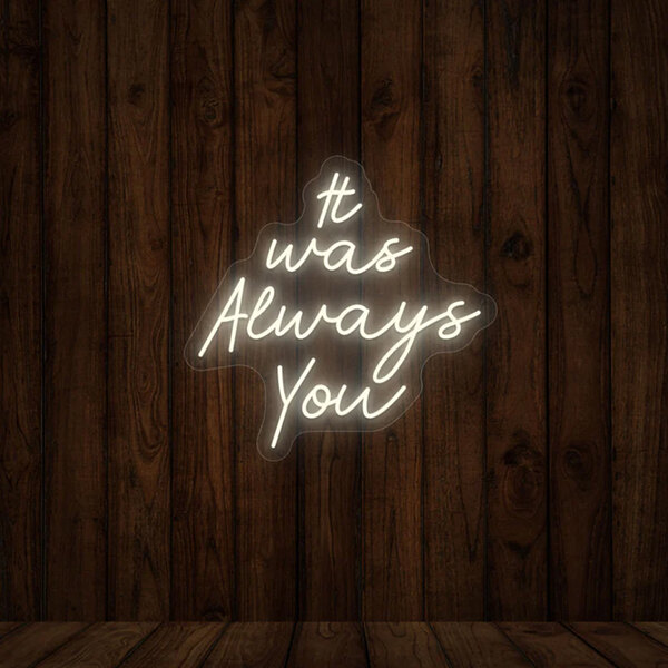 A warm white "It Was Always You" neon sign on a wood wall.