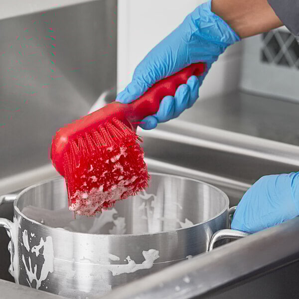 A person in blue gloves using a red Lavex floating utility brush to scrub a pot.