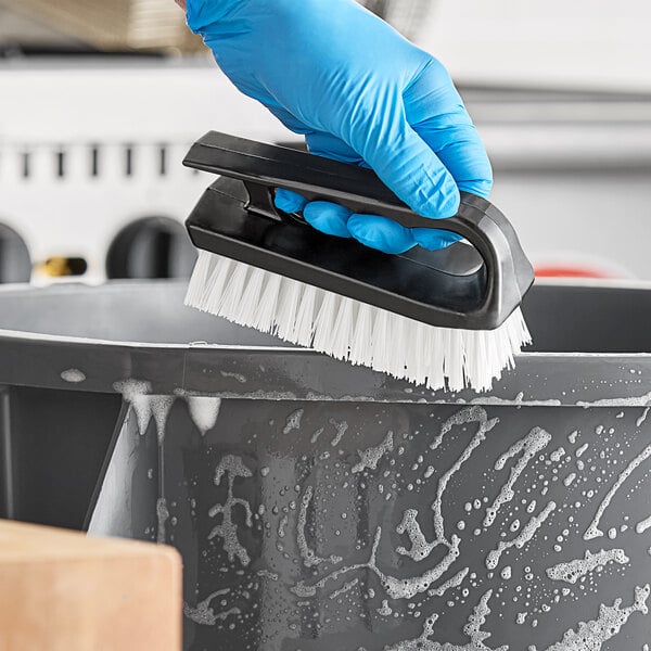 A hand in a blue glove using a Lavex black iron scrub brush to clean a black object.