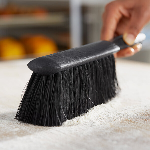 A hand using a black Lavex counter brush to clean up flour on a counter.