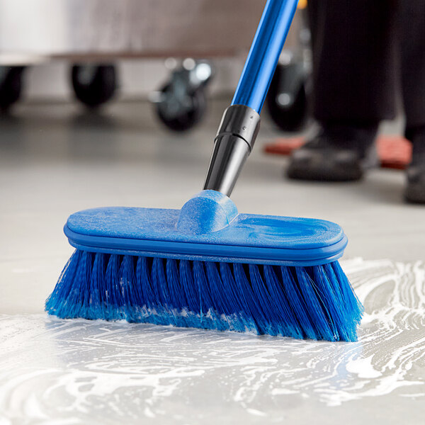 A person using a Lavex blue floor scrub brush with a handle to clean a floor.