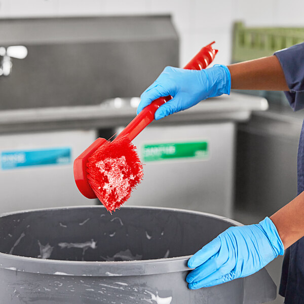A person wearing blue gloves scrubs a bucket with a Lavex red utility brush.