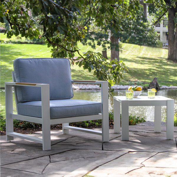 A white square Outdoor Interiors aluminum table on a stone patio with a blue chair.