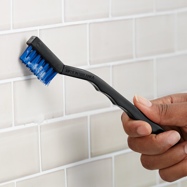 A hand holding a Lavex toothbrush style grout brush with nylon bristles and black handle.