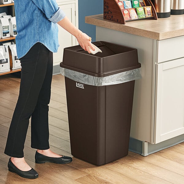 A woman standing next to a Lavex brown square trash can with a plastic bag over it.