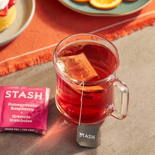 A glass of Stash Pomegranate Raspberry green tea with a tea bag in it on a table with fruit.