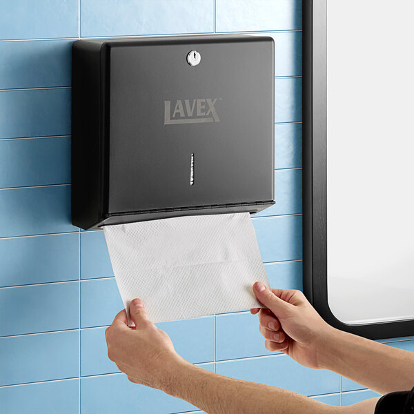 A person using a Lavex Matte Black paper towel dispenser to clean a tile wall.