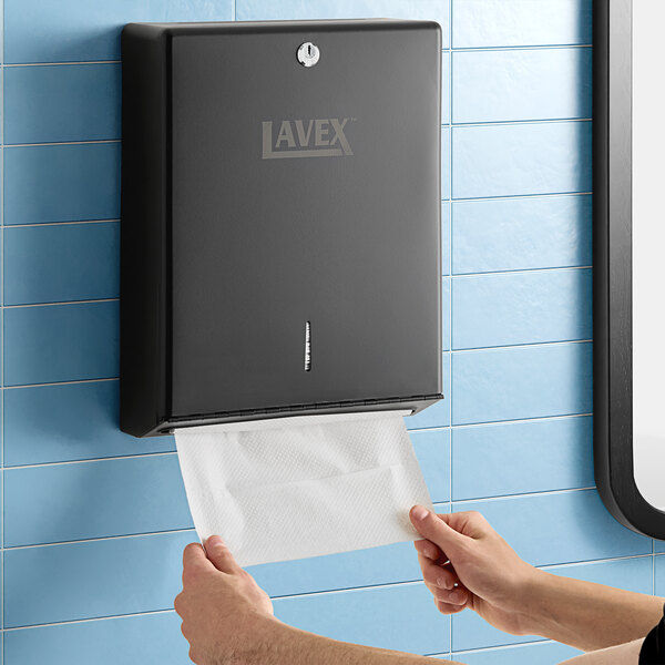 A person holding a paper towel in front of a black Lavex paper towel dispenser.