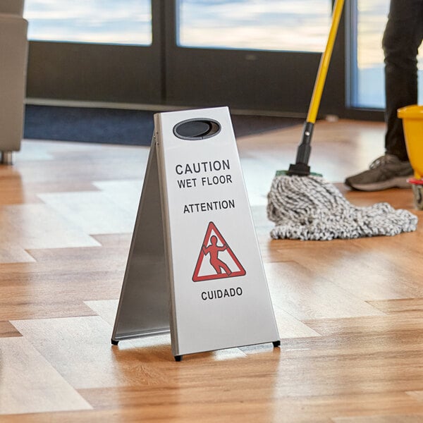 A person using a Lavex stainless steel caution wet floor sign to clean a floor.