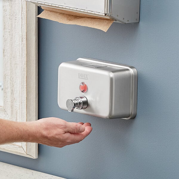 A person using a Lavex stainless steel manual foam soap dispenser on a wall.